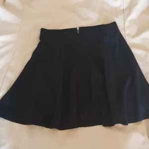 Svart cirkelskuren kjol med dragkedja - En svart, utsvängd kort kjol med silvrig dragkedja bak. Kjolen har en enkel och stilren design som passar perfekt till sneakers eller boots. Materialet känns mjukt och följsamt, och midjan är hög för en snygg siluett. Några enstaka noppor på baksidan (se bilder för skick), därav priset 