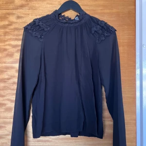Svart blus med spetsdetaljer - Säljer en svart blus med långärmade, lätt transparenta ärmar och hög hals med vacker spets och knappar i nacken. Blusen har dekorativa spetsdetaljer på axlarna och en rak passform. Perfekt för dig som gillar en stilren men ändå romantisk look.