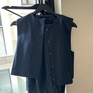 Mörkblå jeansväst i S från Zara och kostymbyxor till i xs - Säljer en stilren mörkblå jeansväst från Zara i rak modell. Västen har tryckknappar framtill och är tillverkad i klassiskt denimtyg. Perfekt att styla över en t-shirt eller skjorta för en cool look.