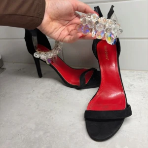 Svarta klackar med kristalldetaljer - Snygga svarta pumps med hög klack och öppen tå. Skorna har en röd innersula och är dekorerade med stora, glittrande kristaller över vristen. Perfekta för dig som vill sticka ut och addera lite glamour till outfiten.