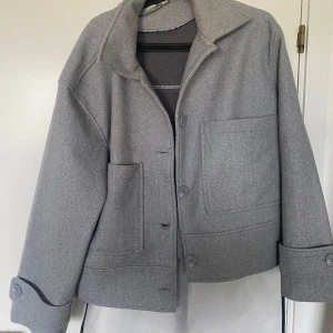 Grå kort kappa från Zink, storlek L - Snygg grå kort kappa från Zink i storlek L. Jackan har stora fickor framtill, bred krage och knappar längs hela framsidan samt vid ärmsluten. Jackan är helt ny och oanvänd!  Enkel att styla till olika outfits. Ordinarie pris 365
