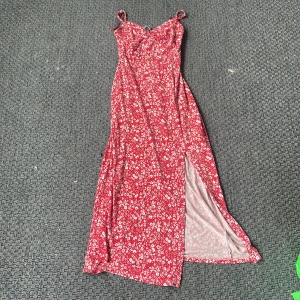 Röd blommig långklänning från Ginatricot - Supersöt röd långklänning från Ginatricot med vita blommor, smala axelband och en snygg slits på sidan. Perfekt för sommarens alla tillfällen! Klänningen har en figurnära passform och är gjord i ett mjukt, lätt material. Tryck ej köp nu