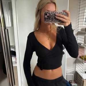 Svart omlottopp med knyt i sidan - Säljer en svart croppad omlottopp med långa ärmar och djup v-ringning. Toppen har knyt i sidan som ger en snygg detalj och sitter tajt över kroppen. Perfekt att styla med höga jeans eller kjol.