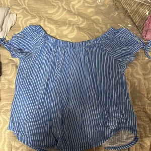 Blåvit randig offshoulder blus - Säljer en blå och vit randig blus med offshoulder-modell och korta ärmar med knyt i sidorna. Luftig och perfekt för varma dagar, toppen har en lös passform och elastisk halsringning. Superfin till jeans eller kjol!