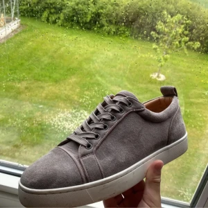 Grå mockasneakers från Louboutin - Snygga grå sneakers i mocka från Christian Louboutin med klassisk röd sula och vita kanter. Skorna har rund tå, snörning och en stilren design som passar perfekt till jeans eller chinos. Riktigt lyxig känsla och ikonisk detalj med den röda undersidan.