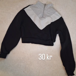 Svart och grå croppad tröja med half zip - Snygg croppad tröja i svart och ljusgrått med coolt V-format mönster och half zip i halsen. Långärmad modell med ribbade muddar och hög krage. Perfekt för en chill och sportig look.