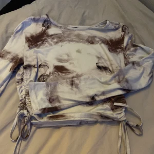 Brun och vit långärmad topp med snörning - Säljer en långärmad topp i vitt med bruna marmorliknande mönster. Toppen har trendig snörning i sidorna som gör att du kan justera passformen och skapa en snygg rynkeffekt. Materialet känns mjukt och stretchigt, perfekt till jeans eller kjol.