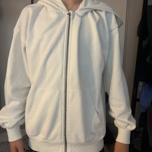 Vit hoodie med dragkedja och huva - Säljer en stilren vit hoodie med dragkedja framtill och stor huva. Jackan har två rymliga fickor och ribbade muddar vid ärmslut och nederkant. Perfekt för chill dagar och enkel att matcha med allt. Oversized fit för en avslappnad vibe.