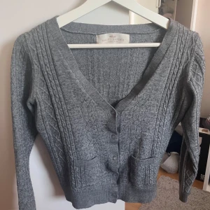 Grå kabelstickad kofta Malou Sander - Grå kabelstickad kofta från Malou Sander med v-ringning, knappar framtill och två små fickor. Perfekt att slänga över en t-shirt eller linne. Storlek S. Inga defekter.
