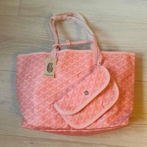 Rosa handväska  - Säljer en helt ny handväska med ikoniskt grafiskt mönster och matchande liten clutch. Väskan har ljusa handtag och detaljer i skinn samt rymligt ljust innerfoder. Perfekt för dig som vill ha en snygg och praktisk väska med lyxig känsla. Hör av er vid fundering!👍🏼🙏🏻