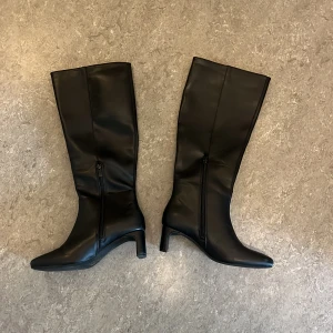 Svarta stövlar med klack och spetsig tå - Snygga svarta stövlar i skinnimitation med spetsig tå och medelklack. Stövlarna har dragkedja på insidan för enkel påtagning och en stilren, minimalistisk look som passar till kjol eller klänning. Perfekta för dig som vill ha en trendig och elegant siluett.