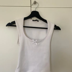 Vit linnetopp med rosett från Zara - Supersöt vit topp från Zara med rundad halsringning och liten rosett framtill. Materialet är mjukt och ribbat,