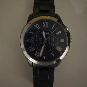 Svart och silver klocka från Fossil - Stilren klocka från Fossil med svart urtavla och silverfärgade romerska siffror. Boetten är i blankt rostfritt stål och armbandet är svart med länkdesign. Kronograf-funktion och tre små visartavlor på urtavlan. Modell FS5230, vattentät till 5 ATM.