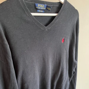 Svart v-ringad tröja från Polo Ralph Lauren - Svart v-ringad tröja från Polo Ralph Lauren med klassisk röd logobrodering på bröstet. Tröjan är i mjuk bomull, har långa ärmar och ribbade muddar vid hals, ärmslut och nederkant. Perfekt för dig som gillar stilrena och tidlösa plagg.