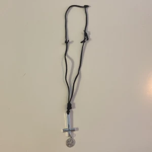 Halsband med omvänt kors och pentagram - Coolt halsband med svart snöre och en silverfärgad hängare i form av ett omvänt kors. Under korset hänger en liten pentagramberlock i samma silverfärgade metall. 