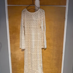 Beige stickad långklänning med knyt - Säljer en beige stickad långklänning med snyggt hålmönster och rund hals. Klänningen har långa ärmar och en fin knytning i nacken som ger extra detalj. Perfekt för dig som gillar boho-vibe och vill ha något unikt i garderoben.