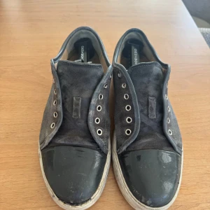 Svartgråa sneakers från Lanvin i mocka - Lanvin sneakers i svart mocka med glansig tå i skinn och platt vit sula. Klassisk rund tå och metallöglor för snörning, men säljs utan skosnören. Snygg kontrast mellan material och färg, perfekt för dig som gillar stilrena och lyxiga sneakers.
