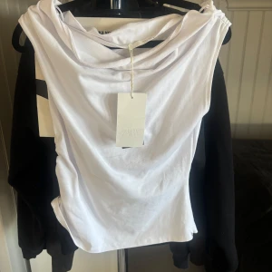 Vit omlott topp från Zara - Snygg vit omlott topp från Zara i storlek S. Toppen har en draperad halsringning och är ärmlös med en cool asymmetrisk design. Tillverkad i mjuk bomull som känns skön mot huden. Perfekt för dig som gillar minimalistisk och trendig stil.