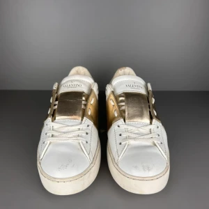 Valentino Garavani Open sneakers vit/guld - Snygga Valentino Garavani Open i vitt skinn med metallic guldpanel över snörningen. Platt sula och rund tå, samt coola nitar på hälen för extra edge. Perfekt för dig som gillar lyxiga detaljer och streetstyle.