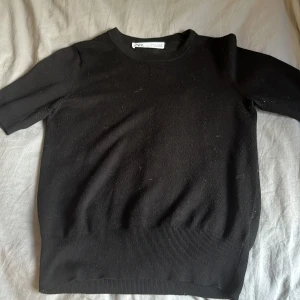 Svart stickad tröja från Zara - En svart stickad tröja från Zara med korta ärmar och rund hals. Tröjan har en ribbad kant nedtill som ger en snygg passform. Perfekt för dig som gillar en enkel och stilren look. 