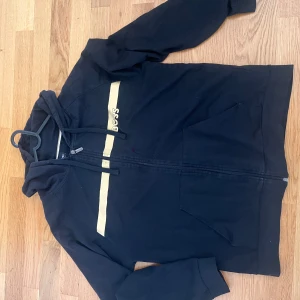 Svart hoodie från BOSS med guldtryck - Svart hoodie från BOSS med dragkedja och huva. Snyggt guldtryck och logga på bröstet, samt två stora fickor framtill. Perfekt för chill dagar och streetstyle. Materialet är mjuk bomull och passformen är normal.