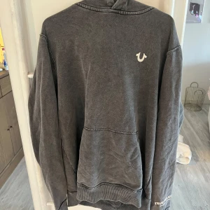 Mörkgrå hoodie från True Religion - Snygg mörkgrå hoodie från True Religion med klassisk logga på bröstet och tryck på ärmen. Tröjan har huva, magficka och ribbade muddar. Perfekt för chill dagar och streetwear-stil. Materialet är mjukt och känns skönt mot huden.