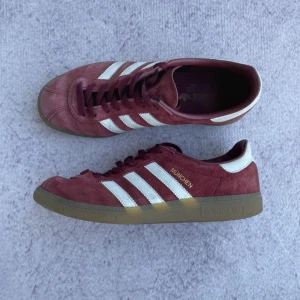 Adidas München vinröda sneakers - Säljer ett par Adidas München sneakers i vinrött mocka med klassiska vita ränder på sidorna och gummisula. Skorna har snörning och rund tå, med guldfärgad text på sidan. Perfekt för dig som gillar retrostil och snygga färgkombinationer.