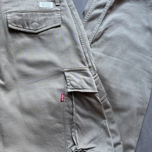 Levi's XX Cargo tapered byxor beige - Säljer ett par Levi's XX Cargo tapered byxor i beige färg. De har klassiska cargofickor på sidorna, knappstängning och bälteshällor. Byxorna är tillverkade i mjuk bomull och har en snygg, avsmalnande passform som ger en chill och modern vibe. Använd 1 gång!