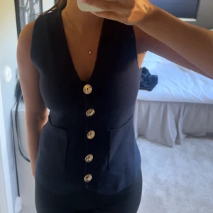 Mörkblå väst med guldknappar - Snygg mörkblå väst med guldknappar. Västen har v-ringad hals och är figursydd.