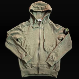 Grön hoodie från Stone Island - Säljer en grön hoodie från Stone Island med klassisk patch på vänster ärm. Tröjan har huva med snörning, dragkedja framtill och två fickor. Materialet är mjuk bomull och passformen är normal. Perfekt för chill dagar och streetwear-stil.