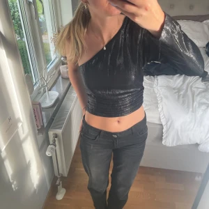 Svart glittrig oneshoulder croppad topp - Snygg svart glittrig topp med oneshoulder och lång ärm. Croppad modell med ribbade detaljer vid midjan.