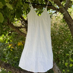 Penny Dress silver med smala axelband - Säljer en vit Penny dress från Fira i storlek L med silverfärgade glittriga ränder. Klänningen har smala justerbara axelband, v-ringad fram och bak, och är kort i modellen. Materialet är lätt och luftigt, perfekt för varma dagar. Snygg och enkel att styla med accessoarer. Helt ny med tags/ prislapp fortfarande kvar. 