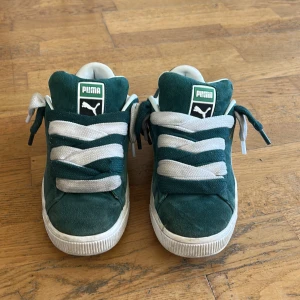 Gröna Puma sneakers i mocka, strl 40 - Säljer ett par gröna Puma sneakers i mocka med vita detaljer och breda vita snören. Skorna har rund tå, platt sula och klassisk Puma-logga i guld på hälen. Insidan är fodrad i vitt och innersulan har ett grafiskt Puma-mönster. Perfekt för dig som gillar retrostil.