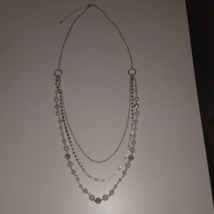 Silverfärgat halsband med pärlor - Snyggt halsband i silverfärg med tre lager kedjor, dekorerat med vita pärlor och glittrande stenar. Perfekt för att lyfta vilken outfit som helst med en elegant touch. Justerbar längd för bästa passform. PRIS KAN DISKUTERAS!