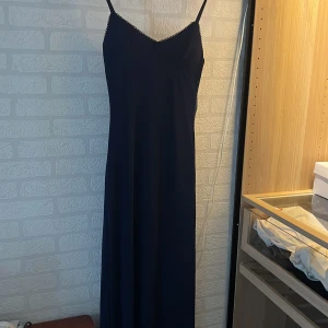 Marinblå långklänning med spetskant - Säljer en stilren marinblå långklänning med smala axelband och v-ringad hals. Klänningen har en fin spetskant längs urringningen som ger en extra detalj. Passformen är normal och tyget känns mjukt och följsamt, perfekt för dig som gillar enkel men elegant stil.