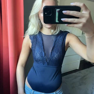Mörkblå spetsig body med mesh - Säljer en mörkblå body med spetsdetaljer och meshpartier både fram och bak. Toppen är ärmlös med v-ringad spets framtill och öppen rygg. Sitter tight och har en snygg, feminin look som passar perfekt till utgång! 