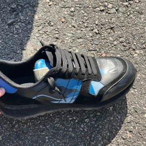 Valentino Garavani blå sneakers - Snygga sneakers från Valentino Garavani med svart och blå camouflage-mönster. Skorna har rund tå, platt sula och svarta snören. Materialmix av skinn och mocka ger en lyxig känsla. Perfekt för dig som vill sticka ut med cool design och exklusivt märke.