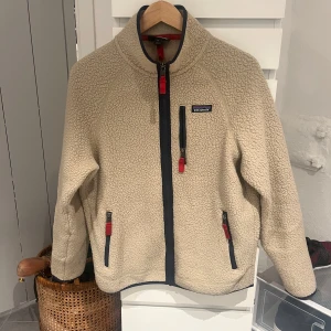 Patagonia fleece Biege - Säljer en beige fleecejacka från Patagonia med mörkblå detaljer och röda dragkedjor. Jackan har hög krage, tre fickor med dragkedja och en mysig teddystruktur. Perfekt för kalla dagar och chill stunder. Snygg och enkel att matcha med jeans eller mjukis.