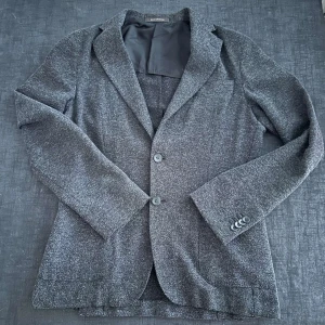 Grå kavaj från Oscar Jacobson - Snygg grå kavaj från Oscar Jacobson med klassisk krage och två knappar framtill. Kavajen har en diskret textur i tyget och fyra dekorativa knappar vid ärmslutet. Perfekt för dig som gillar stilrena och tidlösa plagg. Ny pris runt 4000 kr