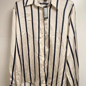 Randig skjorta från Boohoo, strl 34 - Snygg skjorta från Boohoo i storlek 34 med vertikala marinblå ränder på beige botten. Klassisk krage, långa ärmar och knappar framtill. Perfekt för dig som gillar stilrena och trendiga plagg med lite edge.