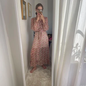 Blommig långklänning med volang - Säljer en romantisk långklänning med blommigt mönster i rosa, grönt och beige. Klänningen har volangdetaljer vid halsen, knappar framtill, knytband i midjan och långa puffärmar. Materialet är lätt och luftigt, perfekt för varma dagar.