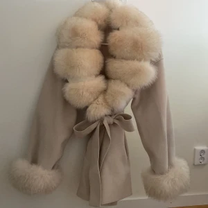Beige kappa med vit ljus brun päls och bälte - Superfin beige kappa med stora fluffiga päls på krage och ärmslut. Knyts i midjan med bälte för en snygg siluett. Perfekt för dig som vill sticka ut och hålla dig varm under höst och vinter. Oversized fit och riktigt lyxig känsla.