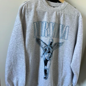 Grå Nirvana sweatshirt  - Säljer en grå sweatshirt från H&M Divided i storlek M med Nirvana-tryck och ängelmotiv på framsidan. Tröjan har rund hals, långa ärmar och är gjord i mjuk bomullsmix. Perfekt för dig som gillar bandmerch och vill ha en chill stil.