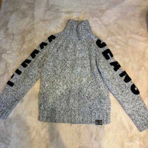 Grå stickad tröja från Pyram Jeans - Säljer en gråmelerad stickad tröja från Pyram Jeans i storlek L. Tröjan har hög krage, långa ärmar med svart text längs båda armarna och ribbade muddar. Materialet är 100% akryl och den har en chill, loose passform som är perfekt för kalla dagar.