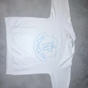 Vit sweatshirt från Moncler med blått tryck - Vit sweatshirt från Moncler med stort blått broderat logotryck på framsidan och klassisk Moncler-patch på ärmen. Tröjan har rund hals, lång ärm och är gjord i mjuk bomull. Perfekt för dig som gillar stilrena och exklusiva märken.