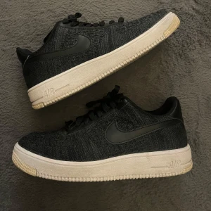 Nike Air Force 1 Flyknit 2.0 (storlek 41) - Svarta Nike Air Force 1 Flyknit 2.0 sneakers med stickad ovandel och klassisk vit sula. Slutsåld modell som ej går att köpa längre, nypris 1899kr säljes för enbart 699kr. Swoosh-logga i svart på sidan, snörning och rund tå. Skorna har en platt sula och är tillverkade i syntetmaterial med stickad textur för extra cool vibe.