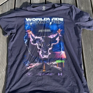 Tränings/gym T-shirt  - Cool mörkblå t-shirt från Under Armour med stor grafisk print framtill med texten 'Worldwide Disruption' och en eldskallig tjur. Snygga lila och blå toner, korta ärmar och rund hals. Materialet är mjuk bomull med mycket stretch, perfekt för dig som gillar streetwear och träning. Mätt kan fås mot efterfrågan