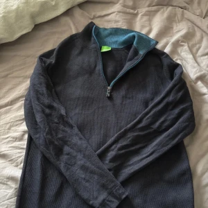Svart half zip tröja från Hugo Boss - Svart långärmad tröja från Hugo Boss med blå detaljer och half zip framtill. Tröjan har ribbad krage med blå insida och broderad blå Hugo Boss-logga på ärmen. Materialet är mjukt och stickat, perfekt för kyliga dagar.