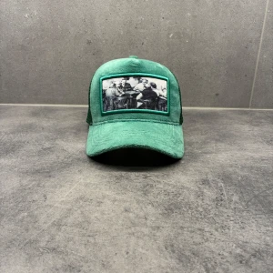 Grön keps Emilio Pendenza  - Snygg grön keps från Emilio Pendenza med mesh på baksidan och ett coolt svartvitt tryck framtill som visar ett gäng personer runt ett bord. Kepsen har böjd skärm och justerbar passform. Perfekt för dig som vill sticka ut med en unik accessoar. 