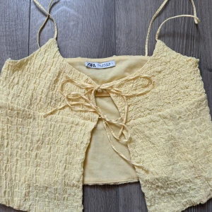 Gul omlott topp från Zara - Supersöt ljusgul omlott topp från Zara med smala axelband och snygg snörning framtill. Toppen har en croppad passform och är gjord i ett mjukt, strukturerat bomullsmaterial som ger en cool textur. Perfekt för varma dagar och sommarvibes!